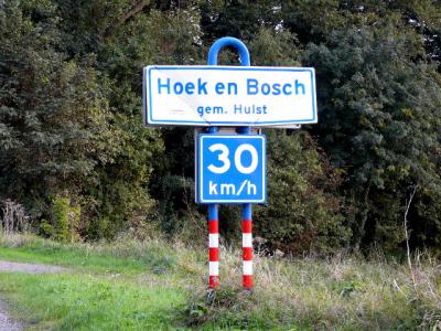 De buurtschap Hoek en Bosch ligt buiten de bebouwde kom en heeft daarom witte plaatsnaamborden, met daaronder borden met een adviessnelheid van maximaal 30 km/uur. De formele maximumsnelheid in dit deel van het buitengebied is 60 km/uur. (© H.W. Fluks)